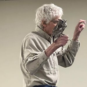 slim man in tan shirt with white hair holds small owl