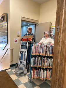 man with empty moving dolly and woman with full book cart stand near elevator