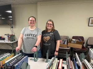 two women smile at the camera with books on tables