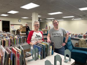 two women smiling in foreground with books on tables while man unpacks boxes in the background