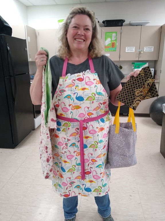 woman in flamingo print apron holds grey flowered tote bag, 2 black and brown clutches, and flowered pillowcase