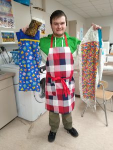 man in red, black, and white checked apron holds Pokémon pillowcase, jellybean print table runner, white and tan tote bag, and black and brown clutch