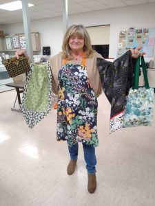 woman with flowered apron holds blue flowered tote bag, shooting star print pillowcase, black and brown clutch, and daisy print table runner