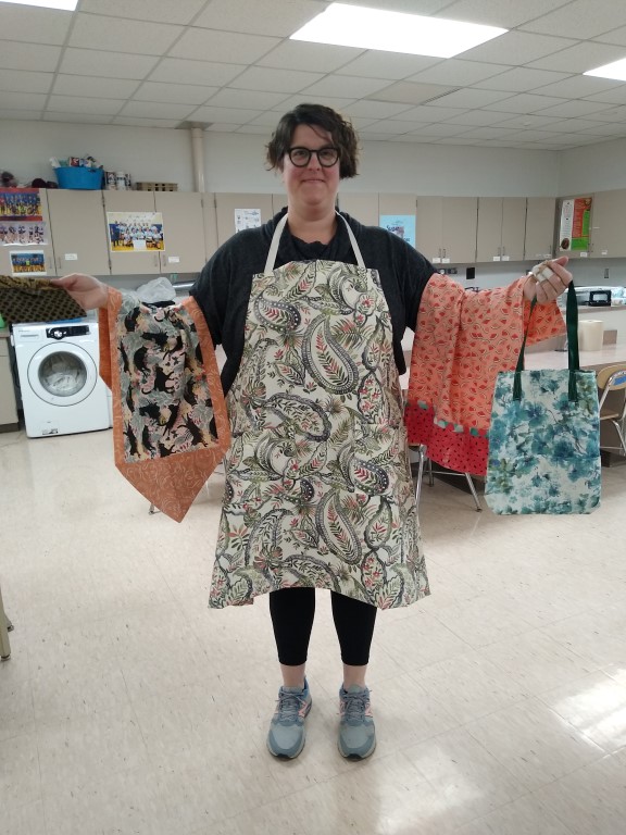 woman with paisley apron holds blue flowered tote bag, watermelon print pillowcase, black and brown clutch, and cat print table runner