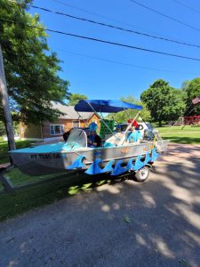 small silver fishing boat with blue roof with three people inside wearing silly fish hats