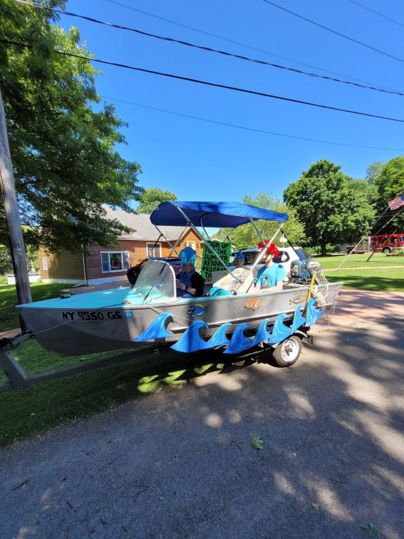 small silver fishing boat with blue roof with three people inside wearing silly fish hats