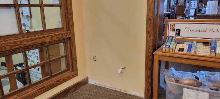section of wall painted beige with pvc and copper pipes sticking out and an uncovered electrical outlet