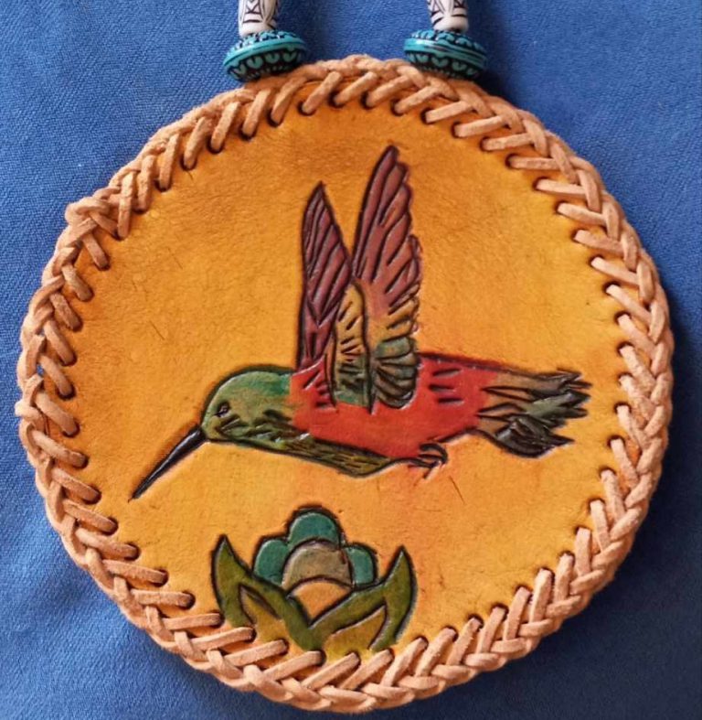 tan circular leather medallion with braided lacing around the rim and image of colorful hummingbird and simple blue flower tooled into it