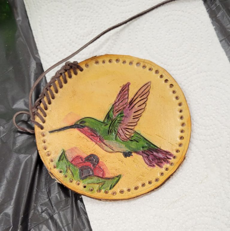 tan circular leather medallion with dark brown braided lacing stitched partway around the rim and image of colorful hummingbird and simple red and purple flower tooled into it