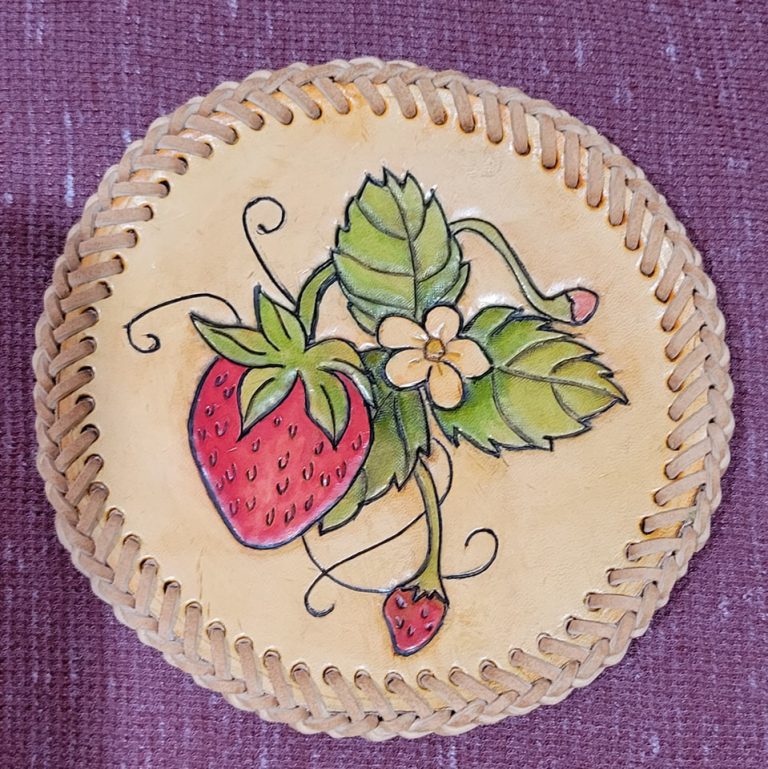 beige circular leather medallion with braided lacing around the rim and image of strawberries, leaves and a strawberry blossom tooled into it