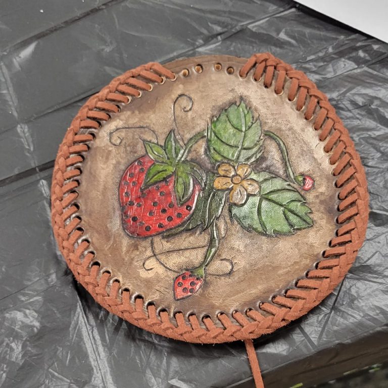 brown circular leather medallion with reddish brown braided lacing around the rim and image of strawberries, leaves and a strawberry blossom tooled into it