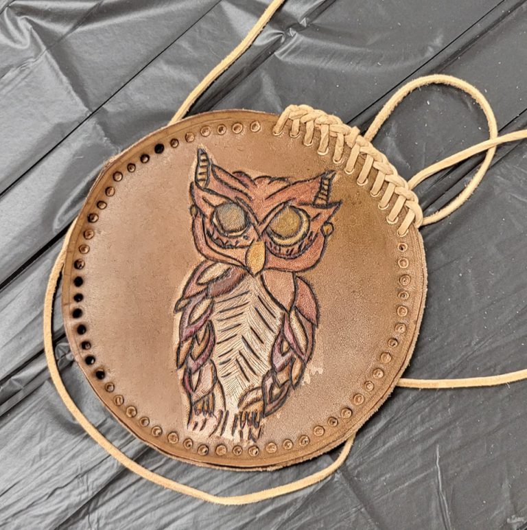 brown circular leather medallion with beige braided lacing being stitched around the rim and image of owl tooled into it