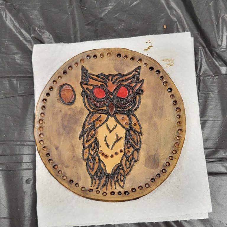 purple-brown circular leather medallion with holes for braided lacing around the rim and image of owl with red eyes tooled into it