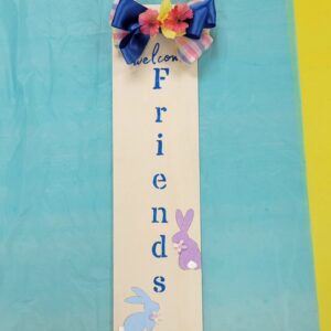 rectangular sign with welcome friends and a purple and a blue bunny painted on and a blue and plaid bow with pink and yellow silk flowers in the center