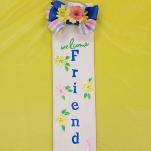 rectangular sign with welcome friends and pink and yellow flowers painted on and a blue and plaid bow with yellow and pink silk flowers