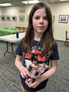 girl with long brown hair and navy shirt with doll with painted face and spiders and skulls glued on