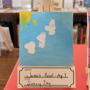 A child’s painting of a sunny day with clouds, displayed on an easel with a handwritten label below.