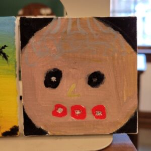A small canvas painting of a simple face with black eyes, light blue hair, yellow nose, and red mouth made up of three dots on a black background.