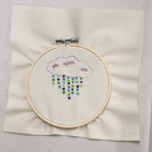 Embroidery of a cloud with colorful beaded raindrops within a hoop.
