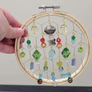 A wooden embroidery hoop with colorful beads hanging inside in a hexagonal wire pattern