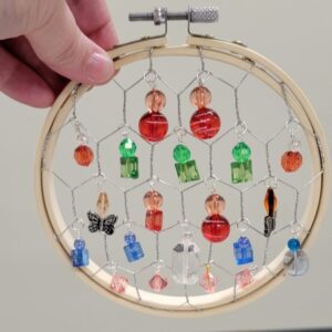 An embroidery hoop with chicken wire, adorned with colorful beads and a butterfly charm, held by a hand