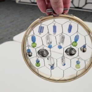 A circular wooden hoop with hexagonal wire, adorned with blue and green beads and silver charms, held by a hand
